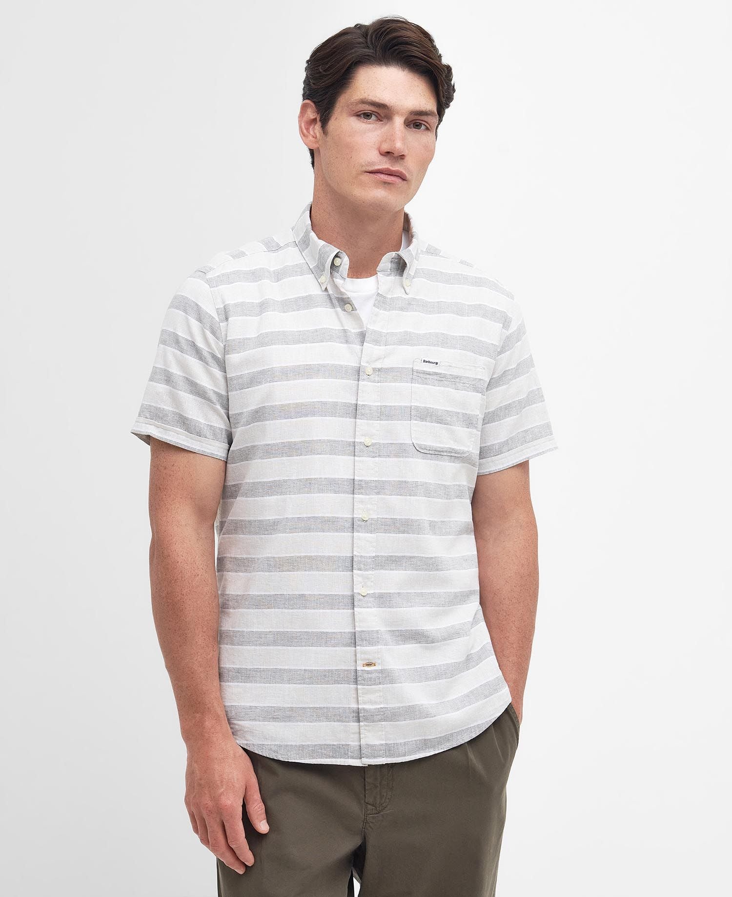 A man wearing a short-sleeved, button-up Barbour Somerby shirt with horizontal stripes in olive green and white, paired with brown trousers.